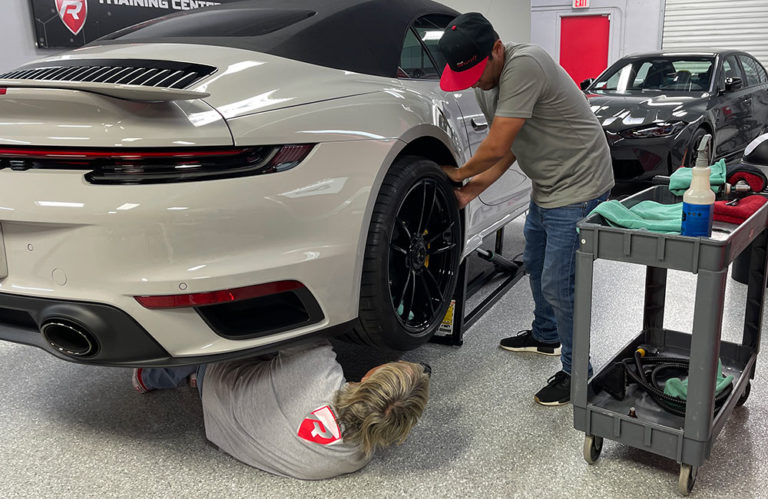 Two people work on a white sports car in the Revivify Coatings Gallery garage. One person is lying on the floor inspecting the underside, while the other stands beside the car, focused on a task around the wheel area. A tool cart with cleaning supplies is nearby, enhancing their efficiency.