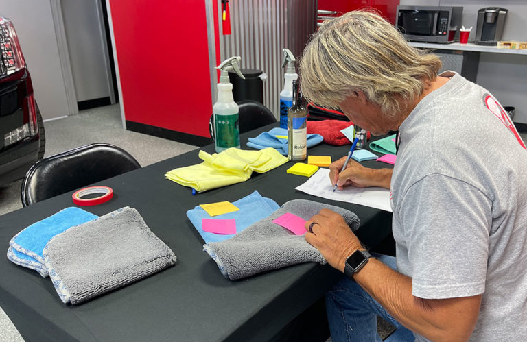 A person with blond hair sits at a table covered with an array of colorful microfiber towels and cleaning supplies, writing on a piece of paper. Sticky notes in different colors are attached to the towels. Behind them, a red and grey wall, cabinets, and a water cooler mark the setting of Revivify Coatings Gallery.