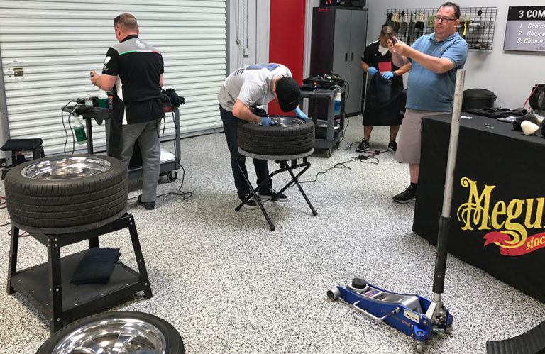 A group of people are working in a garage-like setting. Two individuals are focusing on tire maintenance, with one person cleaning a tire on a stand. Another is using a tool on a tire, while two others appear to be discussing a process at a table labeled "Revivify Coatings.