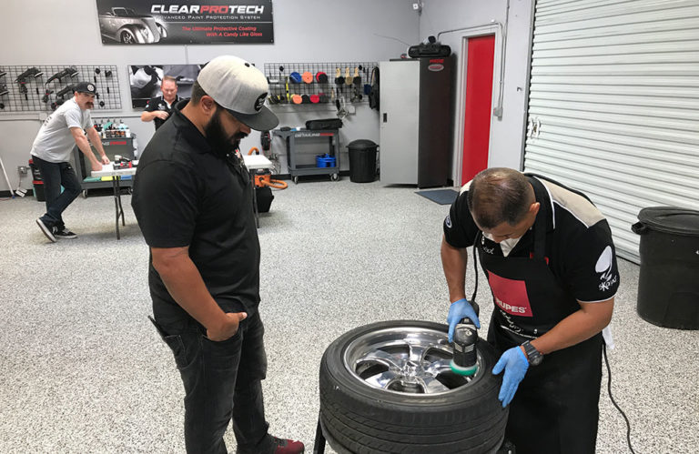 In the Revivify Coatings Gallery, a person is buffing a car wheel with a machine in the garage, while another watches closely. In the background, a small group of people engages in various activities, and an assortment of tools and car care products is organized on a wall-mounted rack.