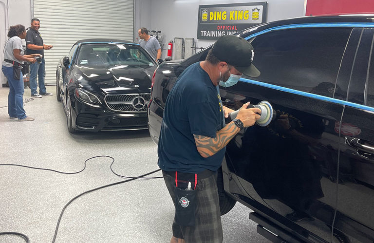 A man wearing a cap and a face mask is using a polishing tool on a black car at the Revivify Coatings Gallery garage. Another man polishes a black Mercedes behind him while two others observe. The garage has signs and equipment in the background.