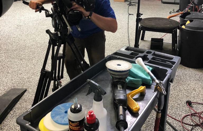 A camera operator films next to a cart filled with car detailing supplies at the Revivify Coatings Gallery. Items on the cart include a buffer, cleaning sprays, microfiber cloths, and various car polish products. The scene appears to be set in a professional auto detailing studio.