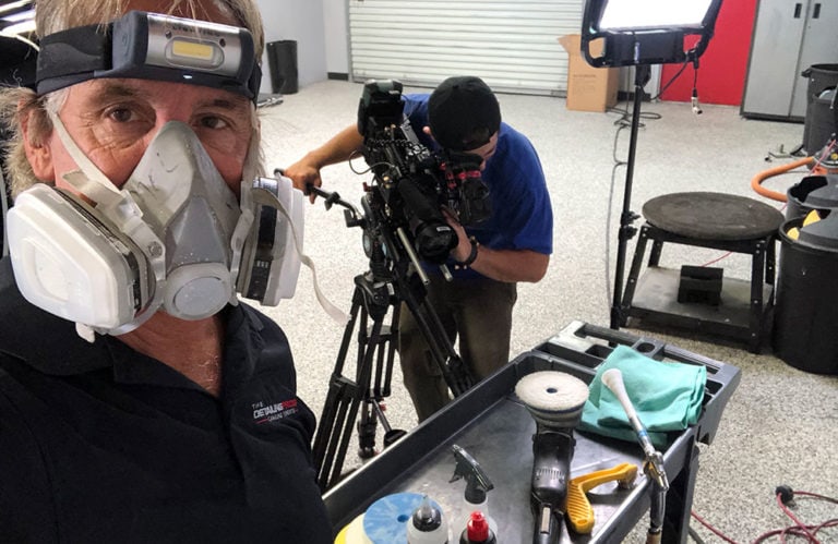 A person wearing a respirator and headlamp stands in a workshop. They are taking a selfie with someone operating a camera in the background. Various tools and cleaning supplies from Revivify Coatings Gallery are on a cart in the foreground, and there are lights set up for filming.