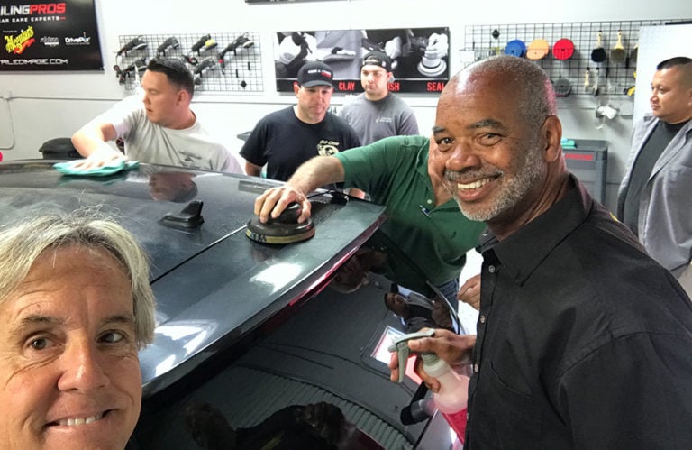 A group of people are detailing a car in the Revivify Coatings Gallery. Several individuals are cleaning and polishing the vehicle, while a man in the foreground smiles at the camera. The workshop features various tools and products on the walls.