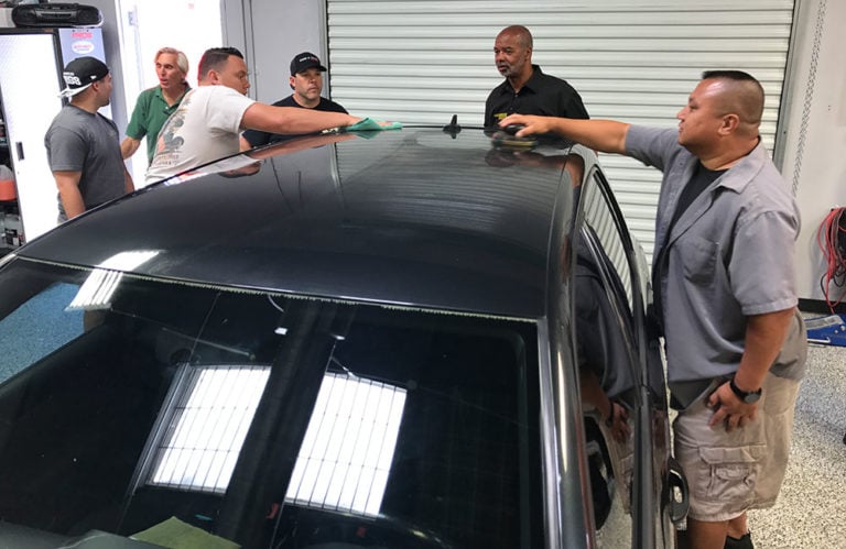 A group of six men are working on a black car inside the Revivify Coatings garage. Some are polishing the car's roof, while others observe and discuss. The background includes a white roller door and various tools and equipment, creating an atmosphere akin to an automotive gallery.