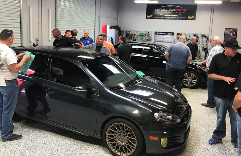 A group of people are gathered in Revivify Coatings' workshop. Several individuals are cleaning and polishing a black car with green cloths. The workshop, known as the Gallery, has large windows, bright lighting, and various car care products on the shelves in the background.