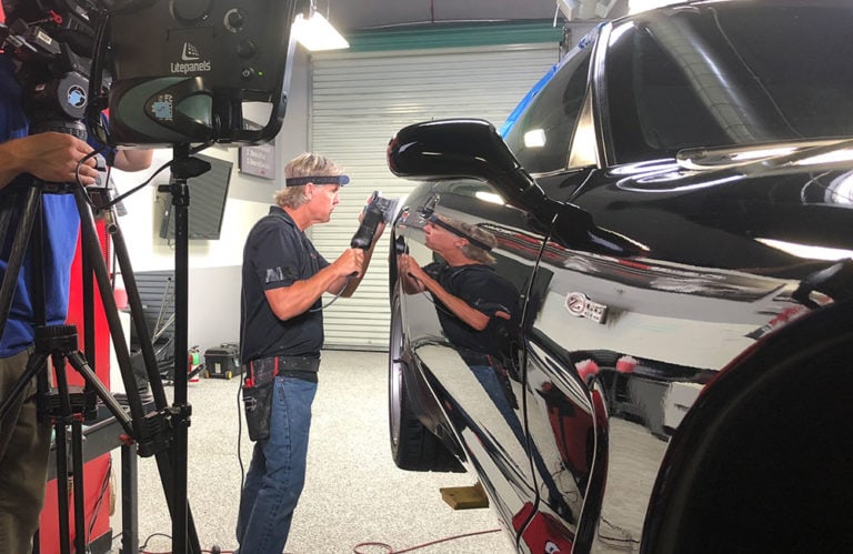 In the Revivify Coatings Gallery, a man closely inspects the side of a black car inside the workshop. Using a handheld tool near the car's surface, he is surrounded by various lighting equipment. The car's glossy paint reflects his image clearly.