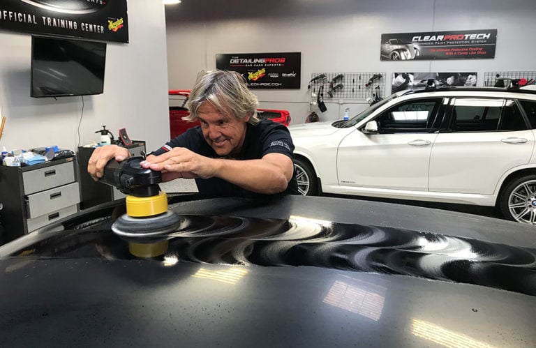 A person polishing the hood of a black car in Revivify Coatings Gallery. They are using a yellow and black power buffer, creating a shiny finish. The workshop has various car care products, including a white SUV, with vibrant posters adorning the walls.