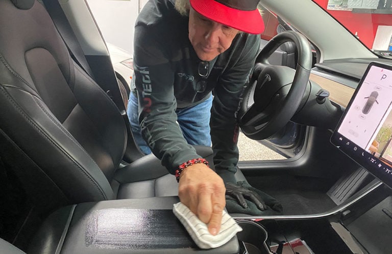 A person wearing a red hat and black gloves is cleaning the inside of a car using a cloth. Their focus is on wiping down the center console area, giving it that signature shine seen in Revivify Coatings Gallery. The car has a large touchscreen dashboard visible on the right side.