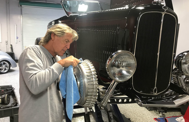 A man with long blonde hair and a grey shirt is polishing a vintage car's headlight using a blue cloth. The car, part of the Revivify Coatings Gallery, has a dark body and an open hood. The setting appears to be a garage or workshop with other classic cars in the background.