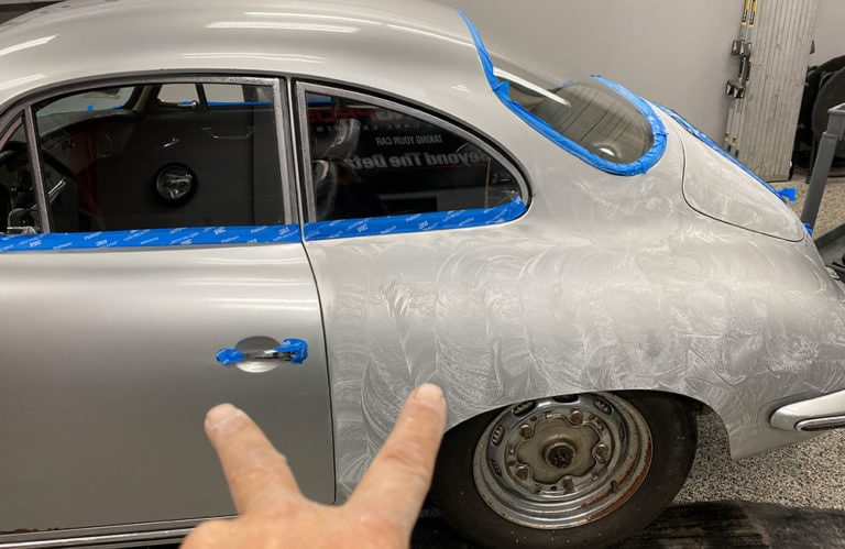 A vintage silver car with blue painter's tape along its edges is undergoing a restoration process. In the foreground, a person points toward the car, with plastic sheeting and various tools visible in the background. This scene could easily belong to the Revivify Coatings Gallery.