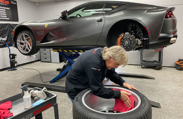 A person cleans a tire with a red cloth in a garage. The tire is detached from a sleek gray sports car, which is elevated on a hydraulic lift. Various tools and cleaning supplies from Revivify Coatings are neatly arranged on a cart beside the person, giving the space an almost gallery-like feel.