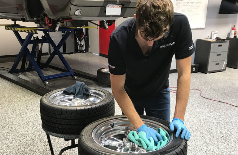 A person wearing a black polo shirt and blue gloves is meticulously polishing a chrome car wheel with a green cloth. Another similar wheel rests on a stand nearby in the Revivify Coatings Gallery. A car is lifted on a hydraulic jack in the background, with a whiteboard and shelves visible in the workspace.