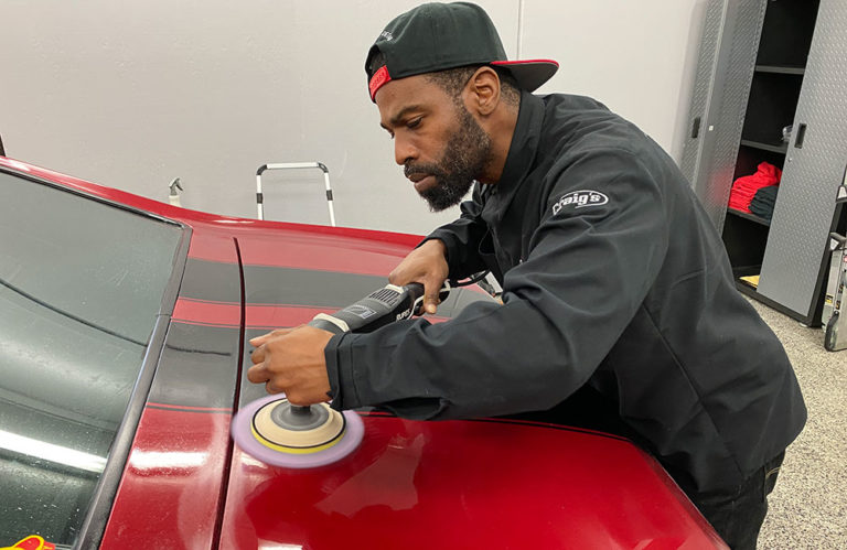 A man wearing a black jacket and a backwards black cap is using a power buffer to polish a red car in the Revivify Coatings Gallery garage. The car's surface is glossy, and the man is focused on his work.