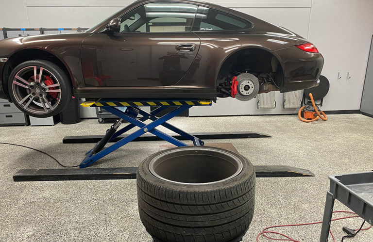 A sports car, elevated on a hydraulic lift, is missing its rear wheel. The removed wheel is placed on the ground in the foreground. The car's rear brake caliper is visible amidst the clean and organized garage environment of Revivify Coatings Gallery.