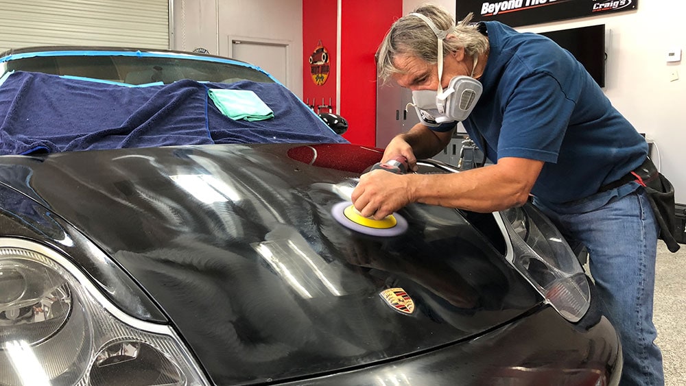 A person wearing a mask and gloves is polishing the hood of a black Porsche with a buffer tool in the Revivify Coatings Gallery. A blue towel and a cleaning cloth are placed on the car's windshield, while various automotive tools are visible in the background.