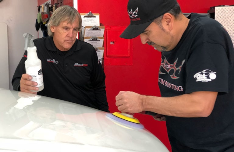 Two men are working on a car in a garage. The man on the left is holding a spray bottle and looking on, while the man on the right is polishing the car's hood with a buffer. Both are dressed in black shirts. Various tools and equipment from the Revivify Coatings Gallery are visible in the background.