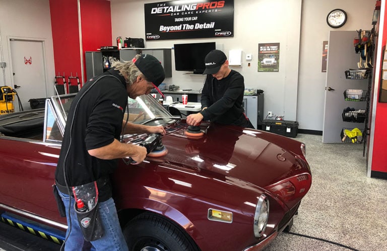 Two mechanics are polishing the hood of a red vintage convertible car inside Revivify Coatings Gallery. Both are wearing black clothing and caps with detailing equipment. The garage, adorned in red, white, and gray decor, boasts detailing supplies and banners promoting top-notch car care.