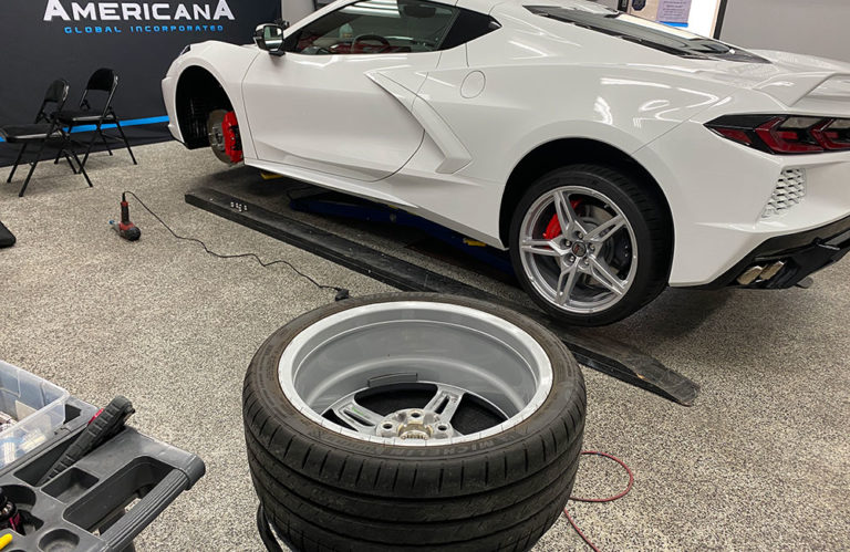 A white sports car is elevated on a lift in the Revivify Coatings Gallery garage. One wheel is removed and rests on the floor in the foreground, exposing red brake calipers. Tools and equipment are scattered around, and a banner in the background reads "Americana Global Incorporated.