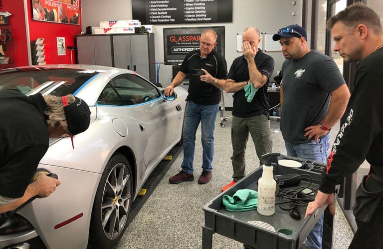 Five men are observing a car in a garage. One man on the left is working on the car, while the others, some holding cloths, focus on different parts of the vehicle. Various tools and cleaning supplies from Revivify Coatings Gallery are scattered on a cart beside the car.