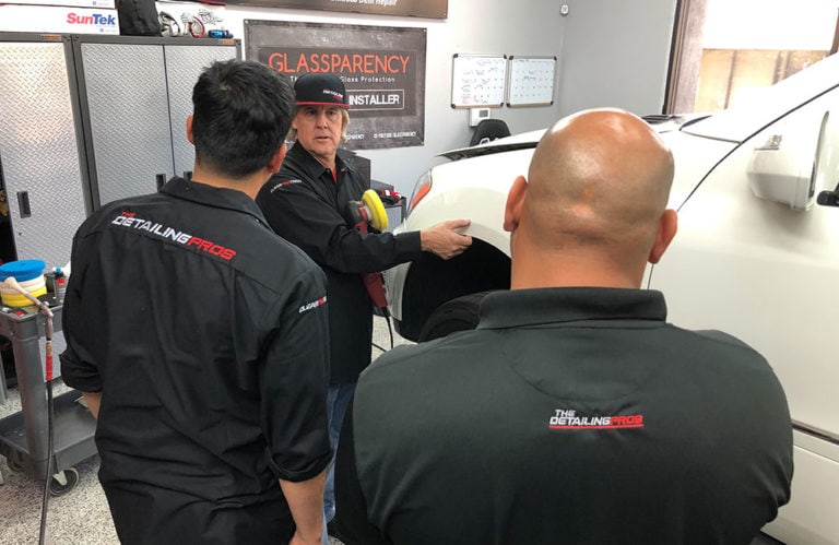 Three men wearing black shirts with "The Detailing Pros" logo are engaged in a discussion in an automotive garage. One man, holding a tool and pointing towards a white vehicle, seems to be explaining or demonstrating something related to car detailing, possibly for the Revivify Coatings Gallery.