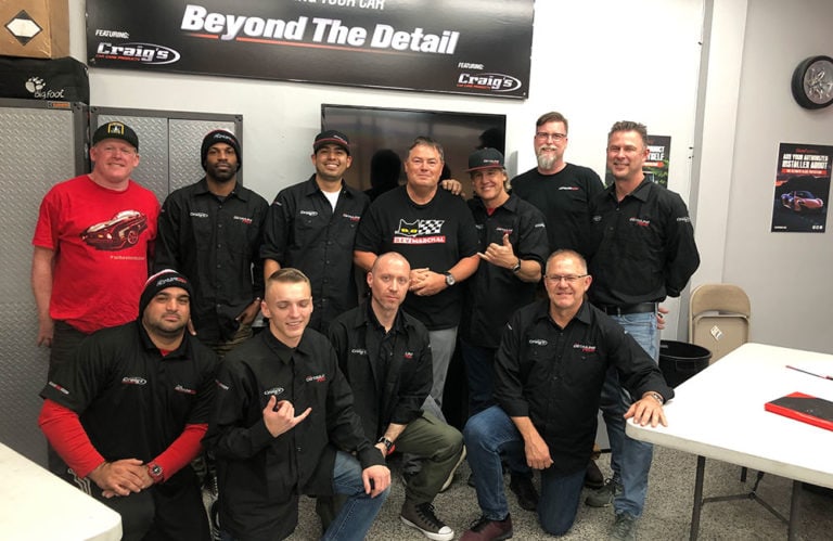 A group of twelve adults, predominantly men, pose together in a garage-like setting. They are wearing black shirts, with one man in a red shirt and hat. A banner above reads "Beyond The Detail." Some are seated on benches while others stand behind them in what appears to be the Revivify Coatings Gallery.