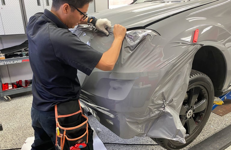 A technician wearing glasses and gloves applies a silver vinyl wrap to the front bumper of a car in the Revivify Coatings Gallery. The technician holds the wrap with one hand while smoothing it with the other. Tools and red toolboxes are visible in the background.