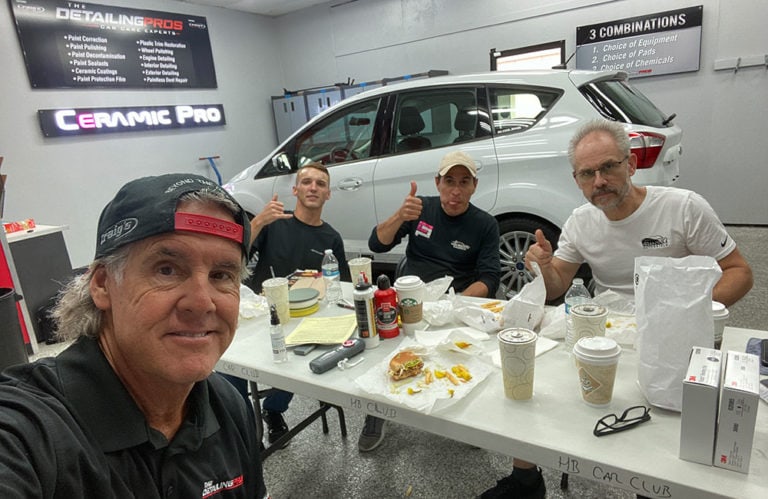 Four men are seated at a table covered with food and drinks, giving thumbs-up gestures. Behind them, in the Revivify Coatings Gallery garage setting, is a white car with signage for Ceramic Pro and The Detailing Pros visible on the walls.