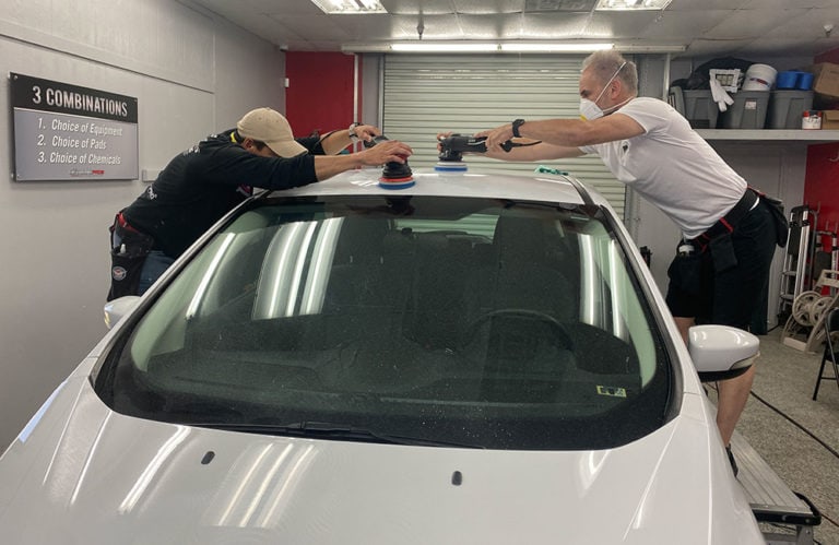 Two men polish the roof of a white car in a workshop. Each holds a polisher, working on opposite sides of the vehicle. Both wear masks, and a sign with equipment instructions is visible on the wall to the left at Revivify Coatings Gallery.