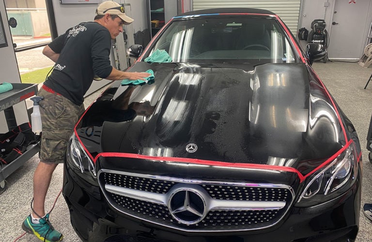 A person wearing a hat and sunglasses is detailing the hood of a black Mercedes inside Revivify Coatings Gallery's well-lit garage. Cleaning supplies and equipment surround the scene as they use a green microfiber towel to polish the car's surface meticulously.