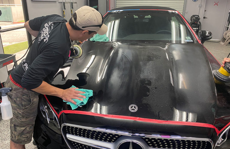 In the Revivify Coatings Gallery, a person wearing a baseball cap and camouflage shorts is polishing the hood of a black Mercedes-Benz in a brightly lit garage. Various tools and equipment are visible in the background while another individual meticulously cleans the hood with a microfiber cloth.