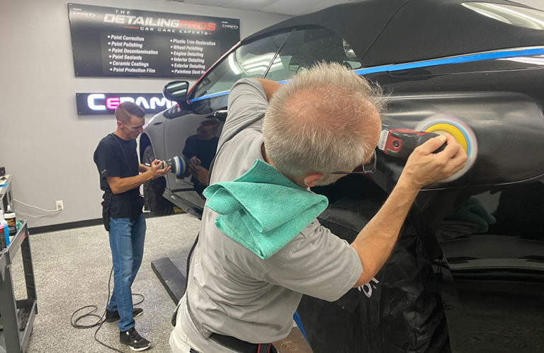 Two people in a professional auto detailing shop are working on a black car. One is buffing the vehicle with a power tool, while the other performs a similar task at the front. In the Revivify Coatings Gallery, a green microfiber towel is draped over the shoulder of the person in the foreground.