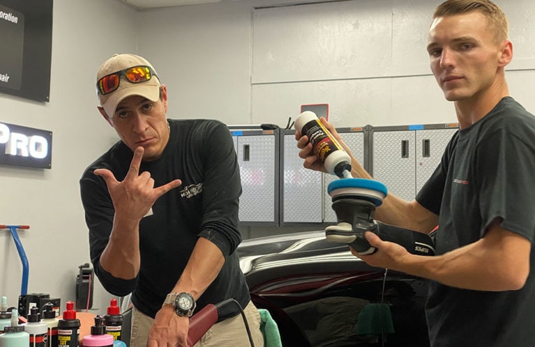 Two men are in the Revivify Coatings Gallery. One is applying polish to a car using a rotary buffer while holding a bottle of polish. The other man is gesturing with his hand. Various detailing products and tools are on a nearby counter, and a car is partially visible in the background.