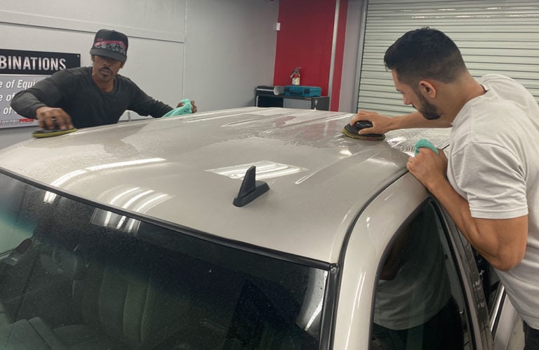 Two men are detailing the roof of a beige car inside the Revivify Coatings Gallery. They use polishing tools and microfiber cloths to clean the surface. A red wall, closed garage door, and equipment can be seen in the background.