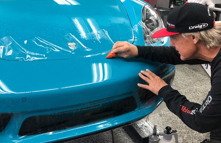 An individual in a black cap and long-sleeved shirt is applying a clear protective film to the hood of a blue sports car using an orange tool in the Revivify Coatings Gallery workshop.