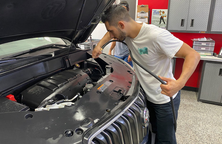 A man in a white t-shirt uses a vacuum hose to clean the engine bay of a car with its hood open. The garage, part of the Revivify Coatings Gallery, is equipped with a red and grey cabinet, a counter with storage, and automotive tools in the background.