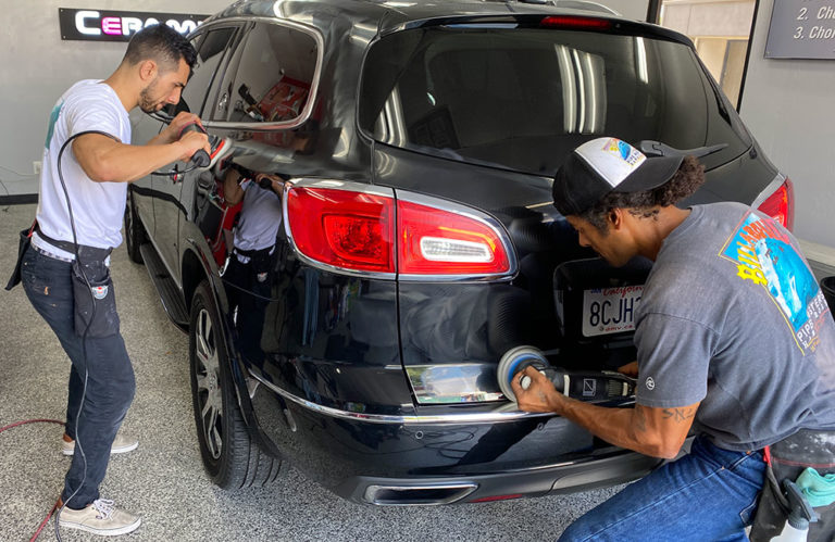 Two people are working on a black SUV in the Revivify Coatings Gallery. One is polishing the rear bumper, while the other is cleaning a rear light. They both have car detailing tools in their hands, and the workshop appears clean and well-organized.