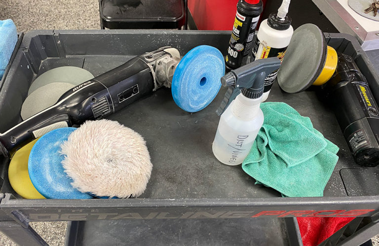 A detailing cart with various car polishing tools and supplies from the Revivify Coatings Gallery. Includes polishing machines, pads, a spray bottle labeled "Distilled Water," green cloth, and bottles of compound on a gray tray with a "Detailing Pros" label.