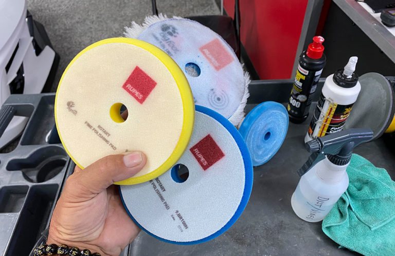 A hand holds two polishing pads labeled "Rupes" in a workspace at the Revivify Coatings Gallery. Various car detailing supplies, including a spray bottle labeled “Dust Master” and different pads, are on a table nearby. The setting suggests a car detailing or polishing operation.