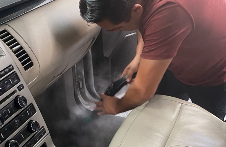 A person in a red shirt is cleaning the interior of a car with a steam cleaner. The image, part of the Revivify Coatings Gallery, focuses on the person's hands as they use the steam cleaner on surfaces near the car's dashboard and a beige seat.