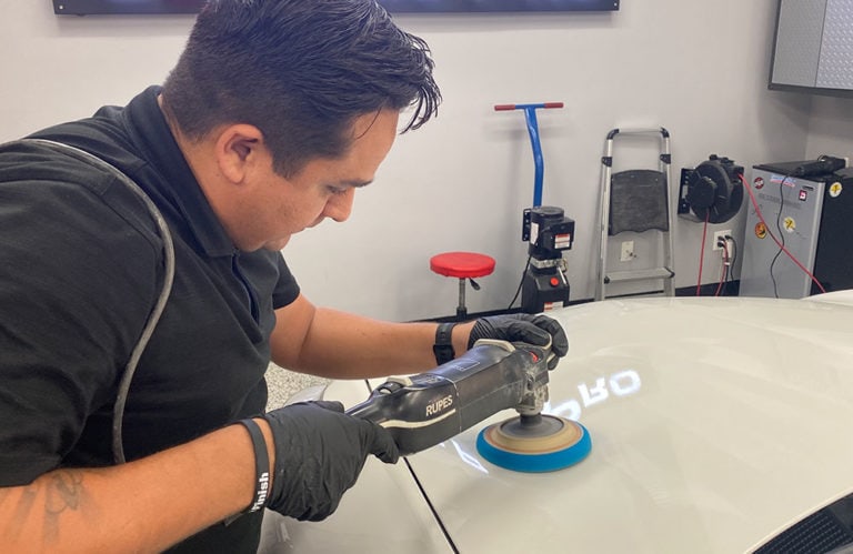 A person wearing black gloves and a black shirt is using a handheld electric buffer to polish the surface of a car in a Revivify Coatings Gallery. Various tools and equipment are visible in the background, including a stool, a wall-mounted power strip, and cleaning supplies.