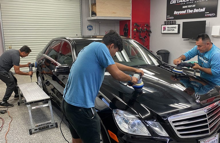 Three people work on polishing a black Mercedes-Benz inside the Revivify Coatings Gallery. Two men use electric buffers on the car's hood and side, while another man polishes the rear door. Various detailing tools and posters are visible in the background, showcasing the shop's dedication to perfection.