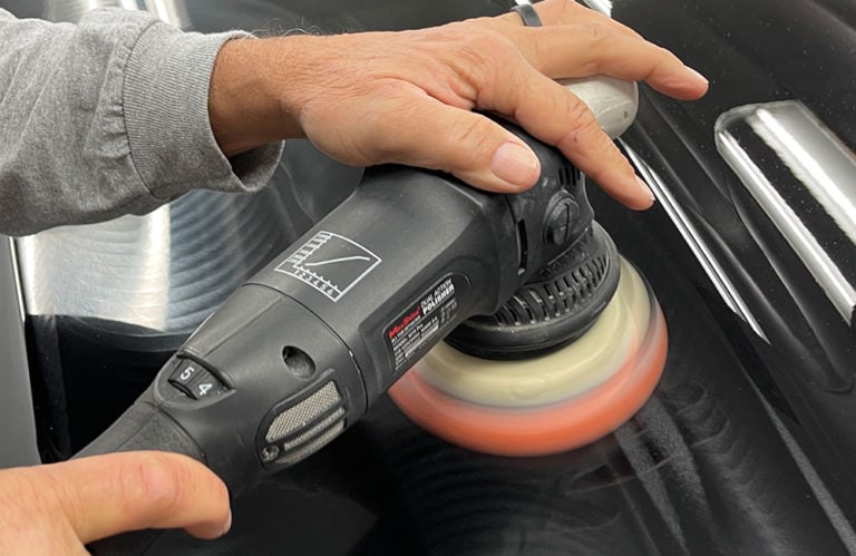 A person is using a power buffer to polish a shiny black car outside the Revivify Coatings Gallery. The machine, showcasing its branded label and red pad, glides effortlessly over the reflective, smooth surface. A wedding ring adorns the person's hand while a grey sleeve peeks from their other wrist.