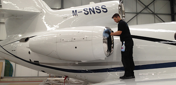 A technician works on the engine of a white aircraft in an indoor hangar. The aircraft has the registration "M-SNSS" painted on its tail section. The technician is wearing black clothing and appears to be focused on the maintenance task.