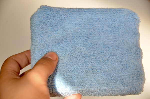 A hand is holding a rectangular, blue Microfiber Sponge against a neutral background. The textured surface of the sponge highlights the typical microfiber material characteristics, making it as versatile as any traditional sponge for cleaning tasks.