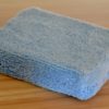 A close-up of a folded, light blue Microfiber Sponge placed on a wooden surface. The sponge has a soft, fuzzy texture and is neatly arranged, displaying its absorbent microfiber fabric.