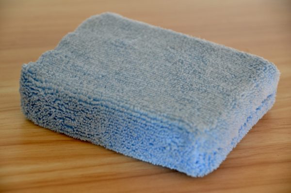 A close-up of a folded, light blue Microfiber Sponge placed on a wooden surface. The sponge has a soft, fuzzy texture and is neatly arranged, displaying its absorbent microfiber fabric.