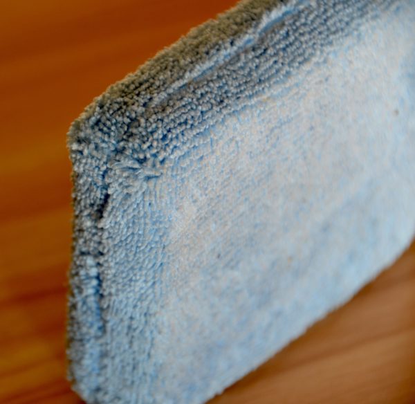 Close-up view of a light blue Microfiber Sponge, standing on its edge, showcasing its soft, textured surface. The sponge is positioned on a wooden surface alongside another cleaning cloth, capturing the contrast between the fabric and the wood grain.