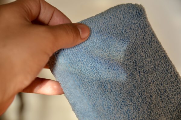 A hand holding a Microfiber Sponge close up. The sponge, with its textured surface, gleams softly under the light, highlighting the intricate fibers. The background is softly blurred, enhancing the focus on the microfiber's detail.