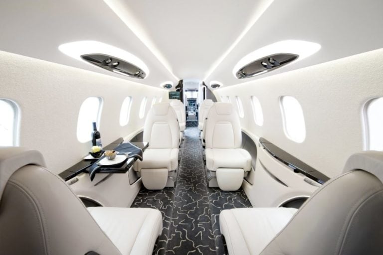 The interior of a luxury private jet featuring plush white seats arranged in pairs along the sides, a patterned carpet, and natural light streaming in from oval windows. A small table with a wine bottle and snacks is visible on the left-hand side.